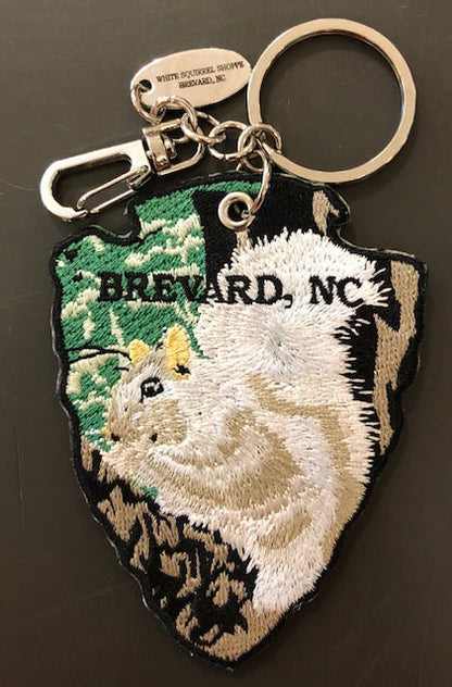 An arrowhead-shaped embroidered key chain with a white squirrel design on this side.Featuring a silver charm with 'White Squirrel Shoppe, Brevard, North Carolina'.