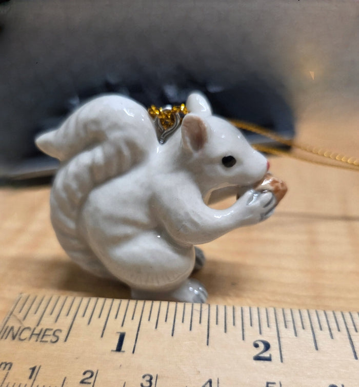A hand painted  porcelain white squirrel ornament with a gold hanging loop, holding an acorn in its mouth close up and size.